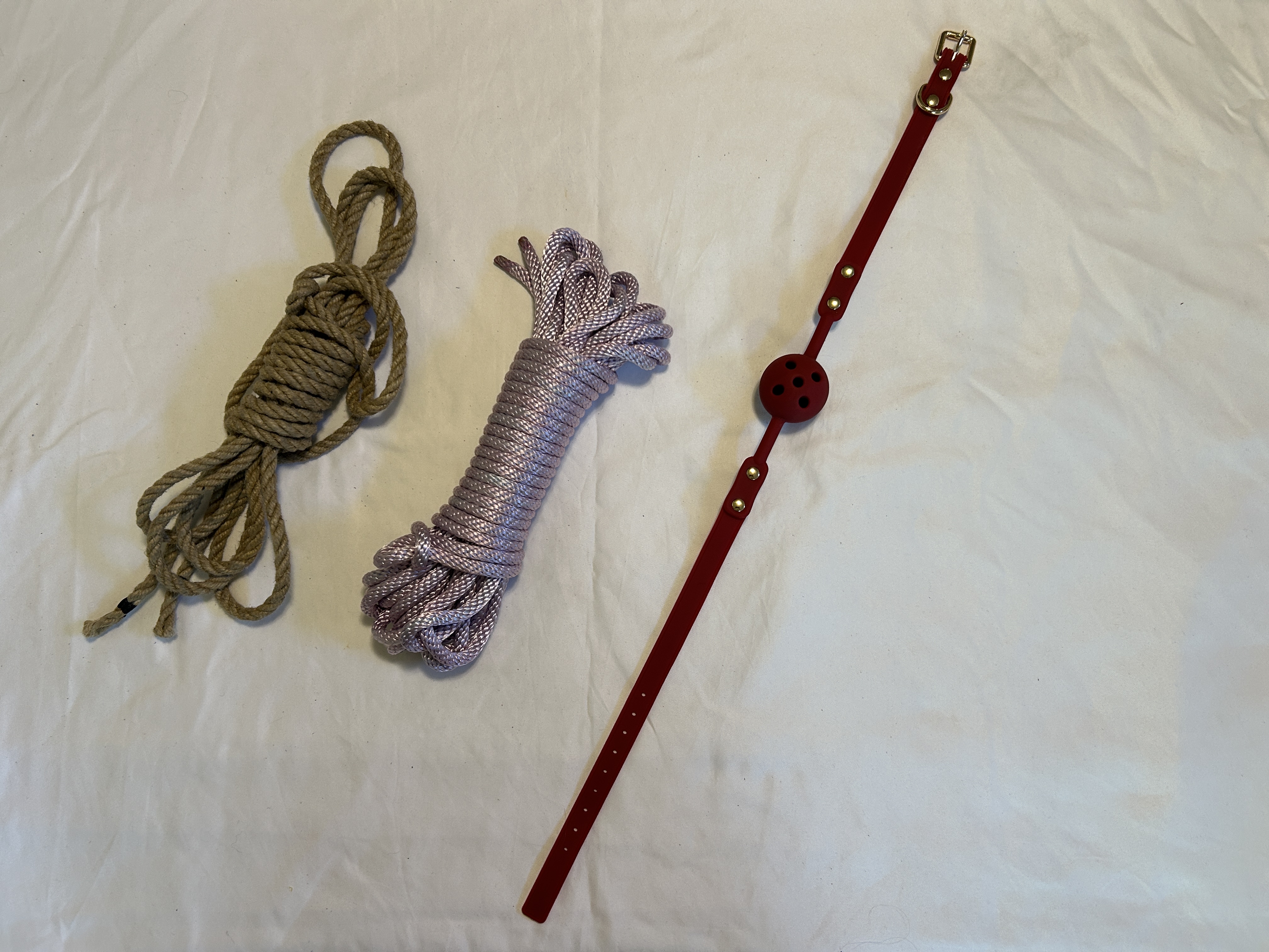 a purplenylon rope, jute rope, and red silicone gag resting on a white sheet